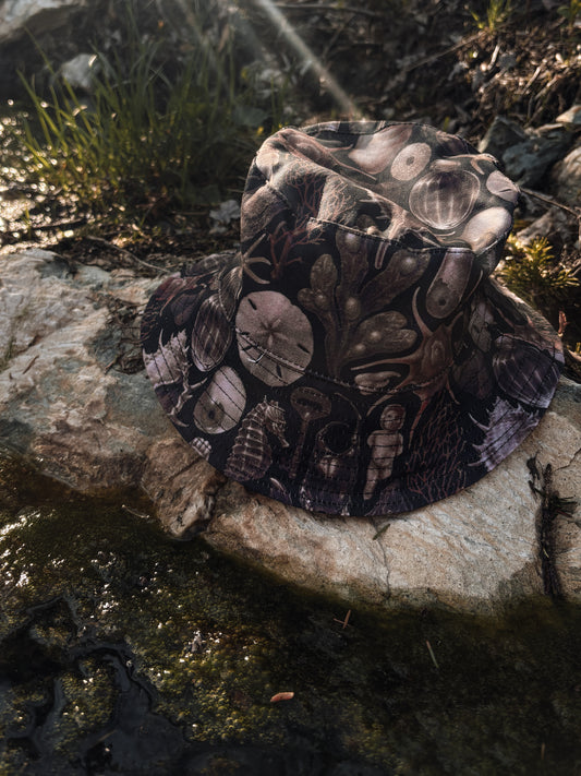 Black bucket hat with a sea-inspired sublimated design combining oddities and seaside treasures. A nod to beach imagery with a goth twist 