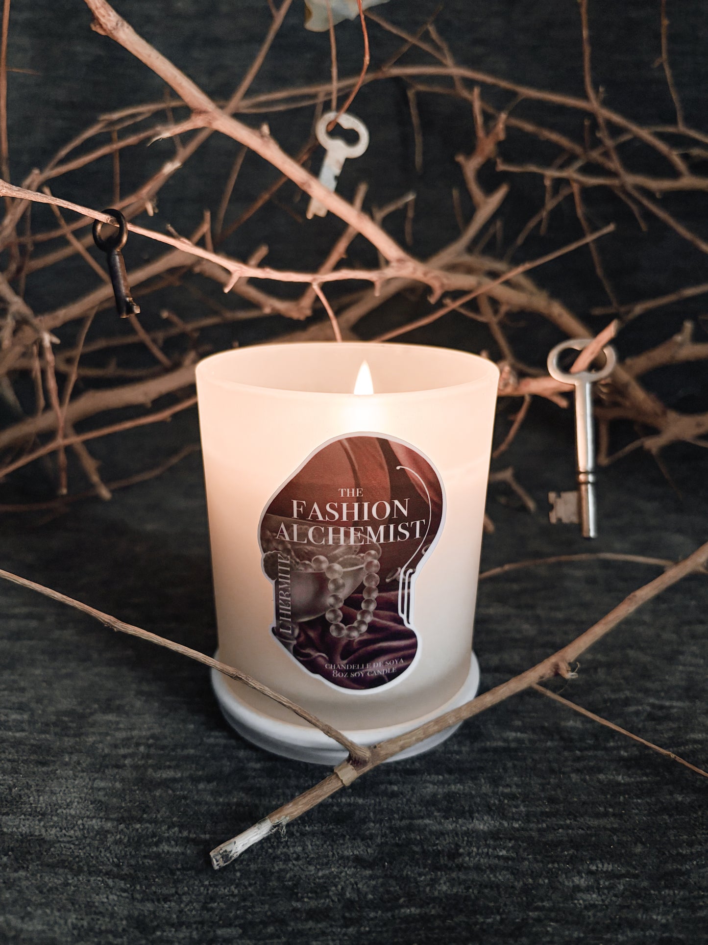 Candle with 'The Fashion Alchemist' label on a dark background with twigs and keys.