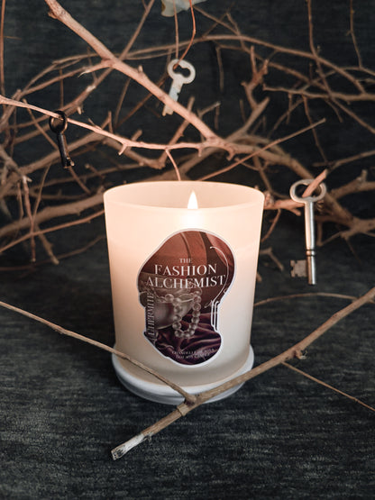Candle with 'The Fashion Alchemist' label on a dark background with twigs and keys.