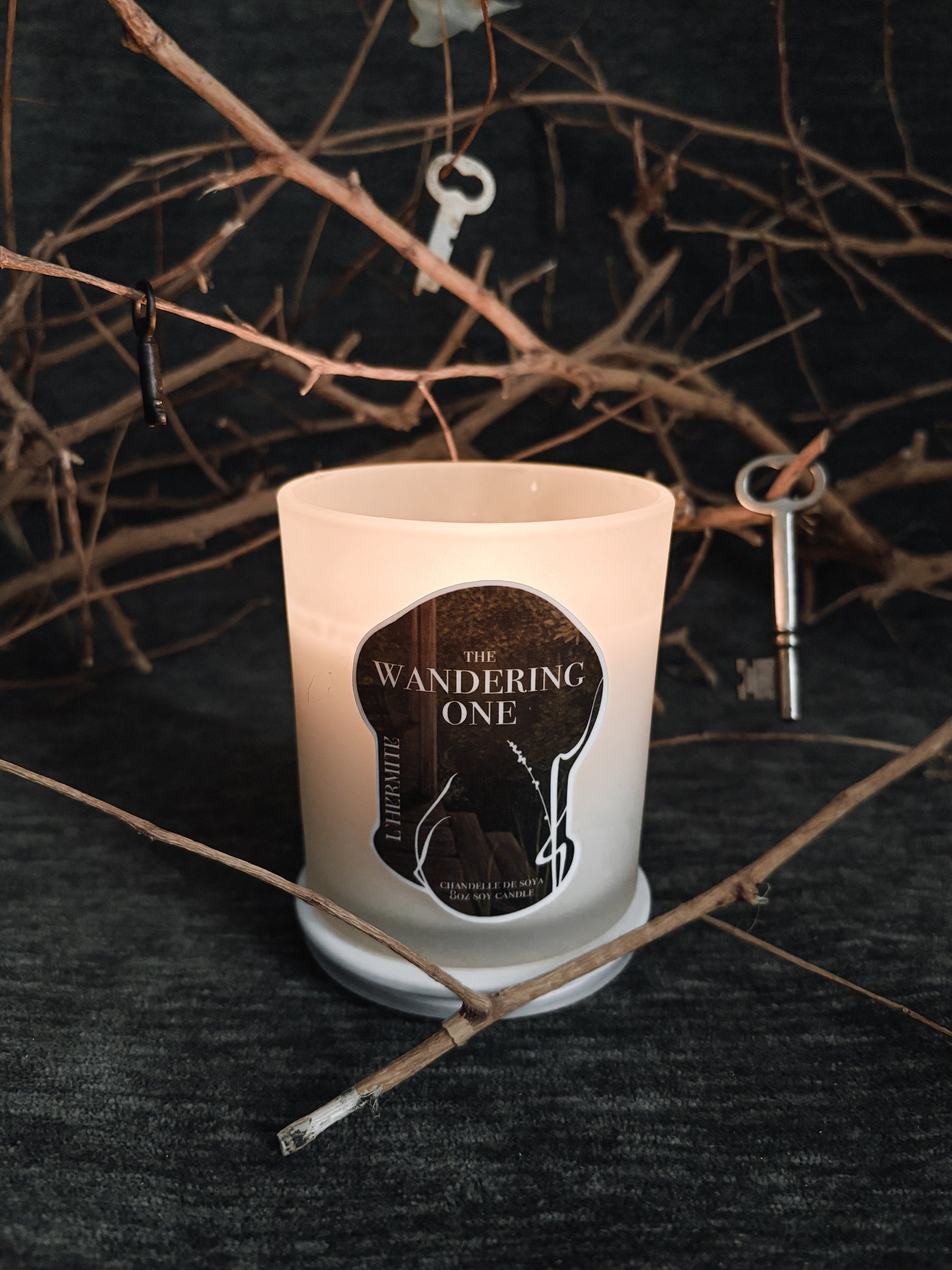 Candle with 'The Wandering One' label on a dark background with twigs and keys.