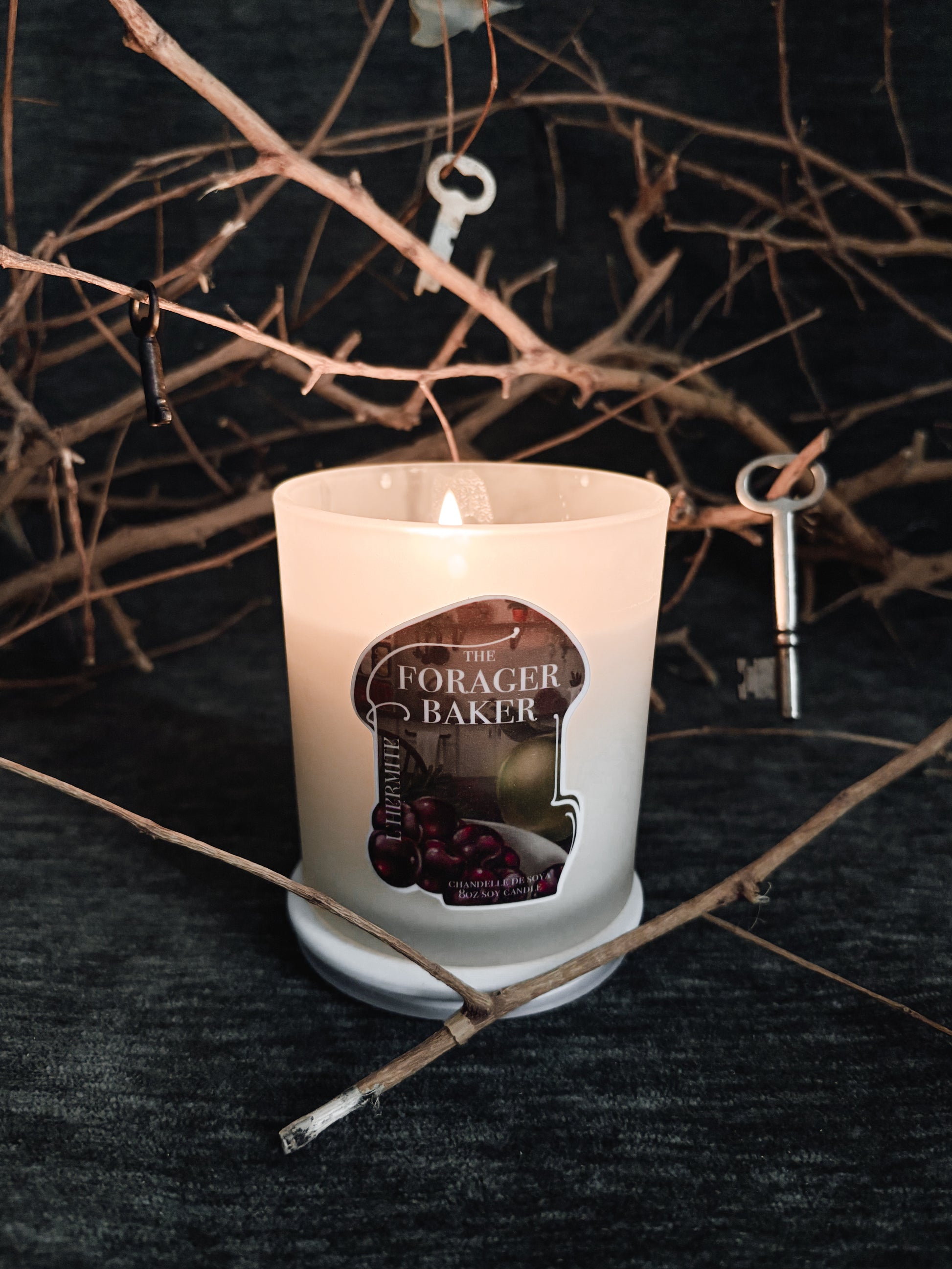 Candle with 'The Forager Baker' label on a dark surface with branches and keys.