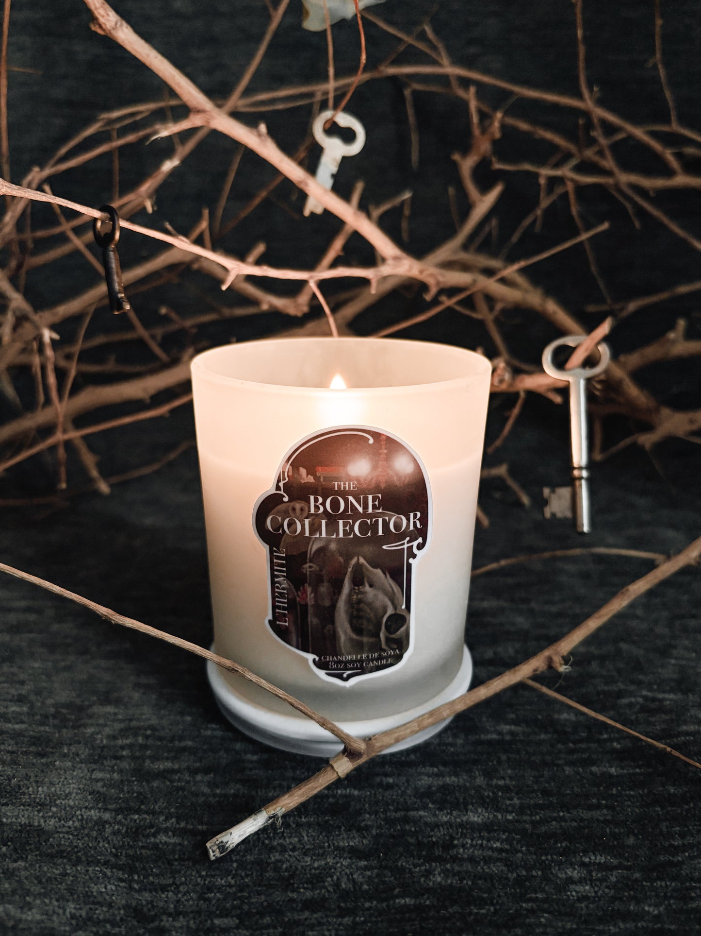 Candle with 'The Bone Collector' label on a dark background with branches and keys.
