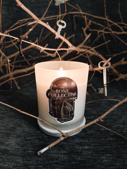Candle with 'The Bone Collector' label on a dark background with branches and keys.